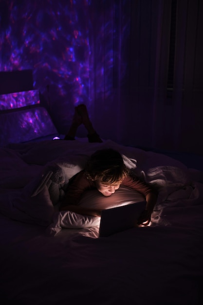 Menino sem sono navegando no tablet enquanto está deitado na cama sob um cobertor no quarto com luz fraca à noite em casa