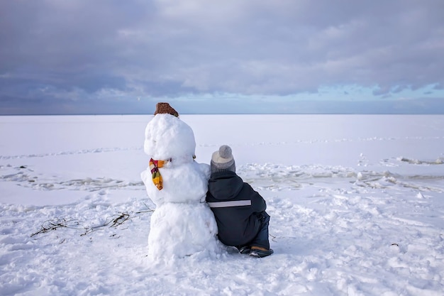 Menino senta-se sozinho com boneco de neve e espera Solidão e amigo feito de neve Clima de inverno