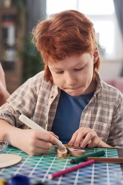 Menino sentado à mesa decorando artesanato com giz de cera durante a aula de arte