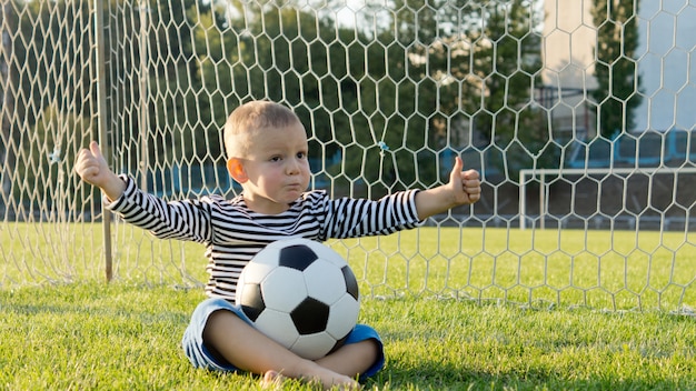 Menino sentado com uma bola de futebol no colo no gol com as redes atrás dele fazendo um gesto de polegar para cima