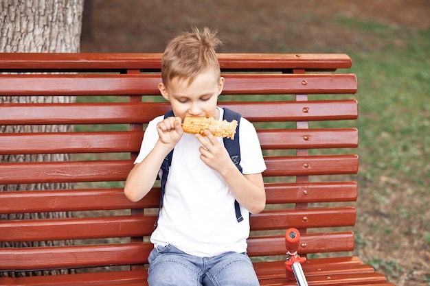 Menino sentado em um banco ao ar livre em um parque e comendo milho fresco cozido.