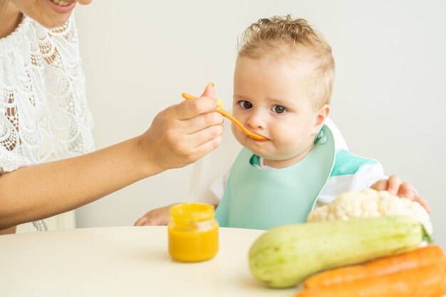 Menino sentado em uma cadeira de childs comendo purê de vegetais na cozinha branca. mamãe alimenta o bebê.
