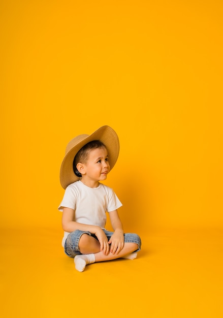 Menino sentado em uma camiseta e shorts com um chapéu de palha e desvia o olhar sobre uma superfície amarela