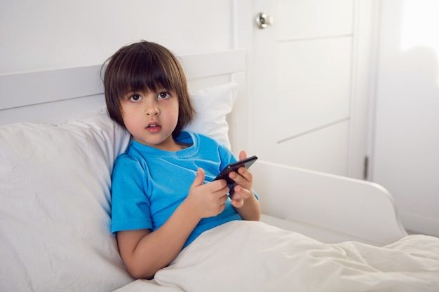 Menino sentado na cama debaixo de um cobertor com um telefone preto e brincando