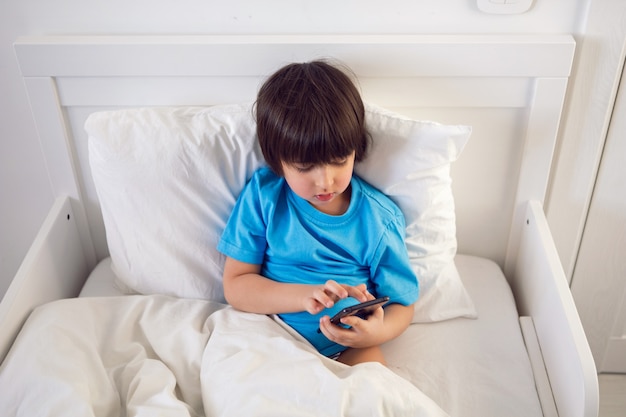 Menino sentado na cama debaixo de um cobertor com um telefone preto e brincando