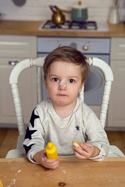 Foto menino sentado na cozinha à mesa com um rolo e farinha no nariz