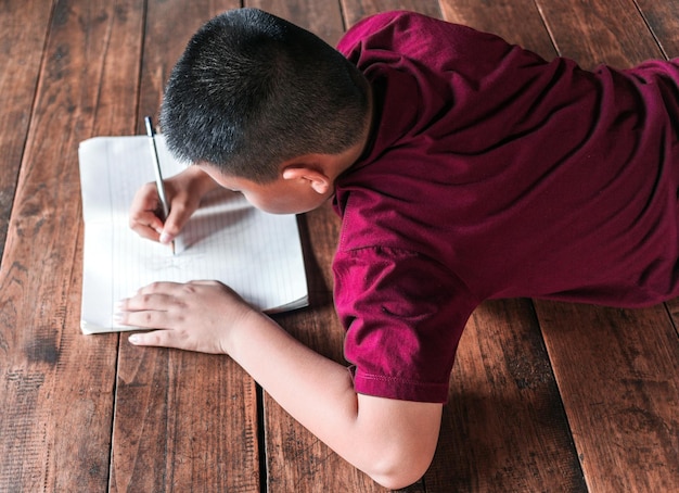 Menino sentado no chão de madeira escrevendo um livro ou fazendo lição de casa em casa Vista superior da escrita infantil