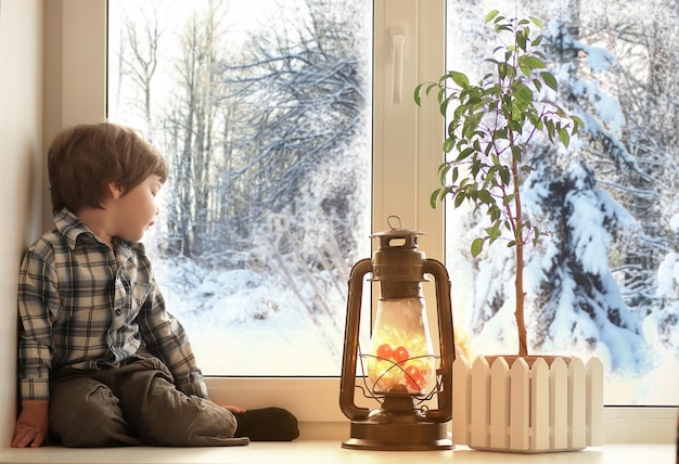 Menino sentado no parapeito de uma janela branca e olhando pela janela para a paisagem de inverno