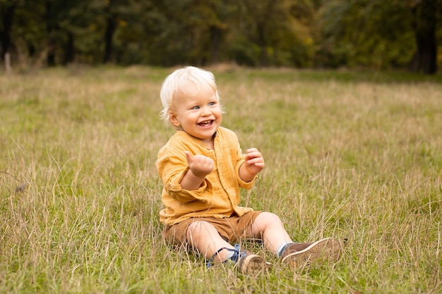 Menino sentado no prado e sorrindo no quente parque de outono com árvores douradas