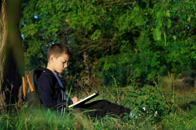 Menino sentado sob uma árvore lê um livro. criança em idade escolar aprende na natureza.