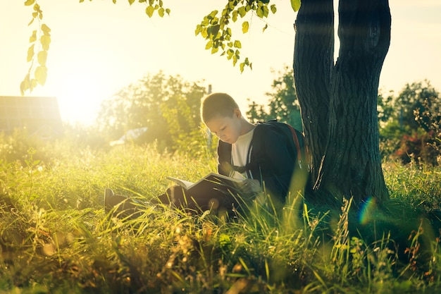 Foto menino sentado sob uma árvore lê um livro. criança em idade escolar aprende na natureza.