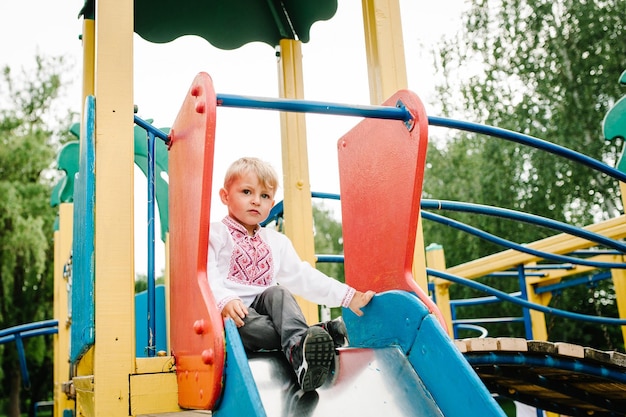 Menino sente-se no escorregador infantil no playground para crianças ao ar livre em uma camisa bordada feche todo o corpo olhando para baixo