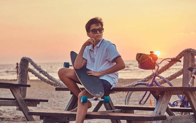 Menino skatista jovem em óculos de sol, vestido de camiseta e shorts, sentado em um banco no contexto de um litoral no pôr do sol brilhante.