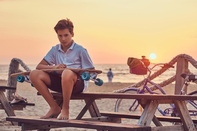 Menino skatista jovem vestido de camiseta e shorts sentado em um banco no contexto de um litoral em um pôr do sol brilhante.