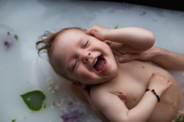 Menino sorridente bonito com olhos fechados toma um banho floral de leite A mãe banha suavemente seu filho pequeno