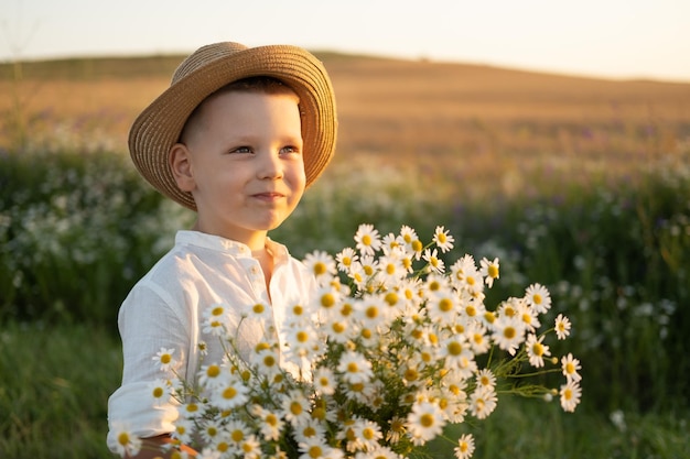 menino sorridente bonito no chapéu de palha detém buquê de camomilas de campo no pôr do sol