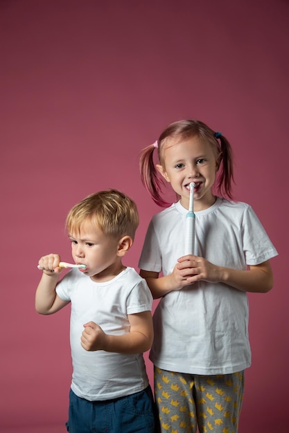 Menino sorridente caucasiano e uma menina limpando os dentes com escova de dentes elétrica sônica e manual em fundo rosa.