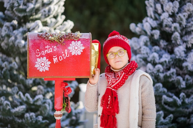 Menino sorridente com chapéu vermelho e óculos verdes com sua carta perto da caixa de correio do papai noel