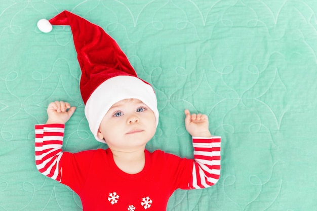 Menino sorridente deitado na colcha verde de Natal bebê com um grande chapéu de Papai Noel vermelho olhando para