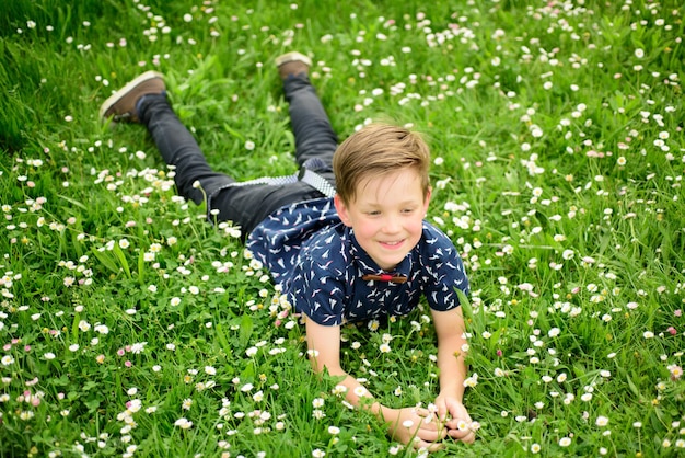 Menino sorridente, deitado na grama, criança fofa, desfrutando no gramado de flores do campo e sonhando