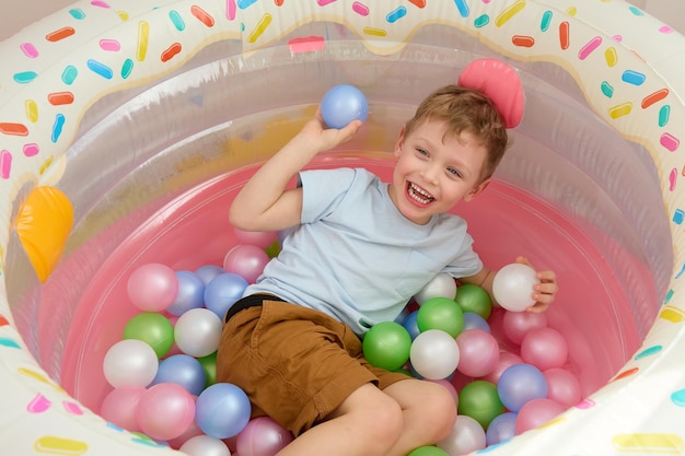 Menino sorridente está se divertindo brincando em casa com balões coloridos Criança caucasiana feliz joga bolas de plástico na piscina seca Vista superior da piscina seca infantil para jogos divertidos