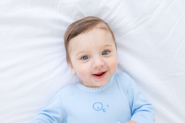 Menino sorridente na cama com um macacão azul, retrato de um bebezinho feliz e saudável