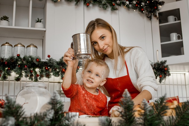 Menino sorridente segura uma peneira de ferro peneira farinha com a ajuda de sua mãe em uma mesa de madeira na cozinha de Natal