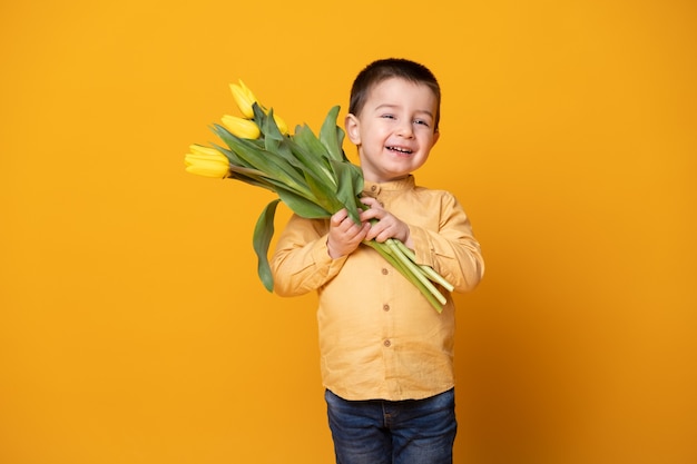 Menino sorridente sobre fundo amarelo do estúdio. criança feliz alegre com buquê de flores de tulipas.