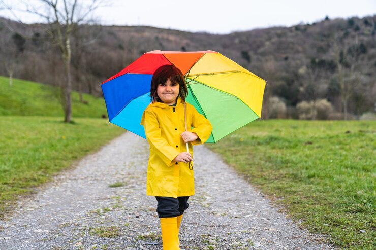 Foto menino sorrindo em um prado ou caminho na floresta, vestindo uma capa de chuva amarela, botas de chuva amarelas e segurando um guarda-chuva colorido de arco-íris na mão