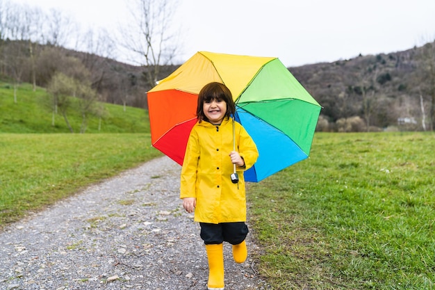 Menino sorrindo em um prado ou caminho na floresta, vestindo uma capa de chuva amarela, botas de chuva amarelas e segurando um guarda-chuva colorido de arco-íris na mão