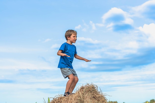 Foto menino sorriso jogar dança careta mostrar suporte de camiseta azul no palheiro fardos de grama seca céu claro dia ensolarado treinamento de equilíbrio conceito infância feliz crianças ao ar livre ar limpo perto da natureza