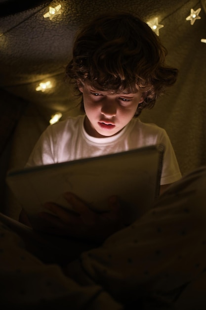 Menino talentoso concentrado desenhando em um caderno de esboços enquanto está sentado sob uma construção de cobertor com guirlandas brilhantes em uma sala escura à noite