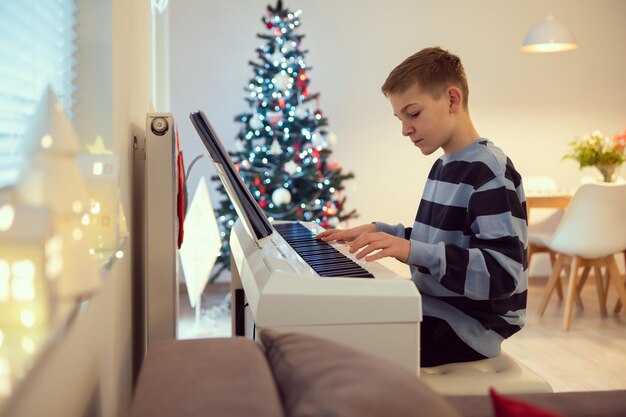 Menino Tennage praticando piano em casa com árvore de ano novo no fundo