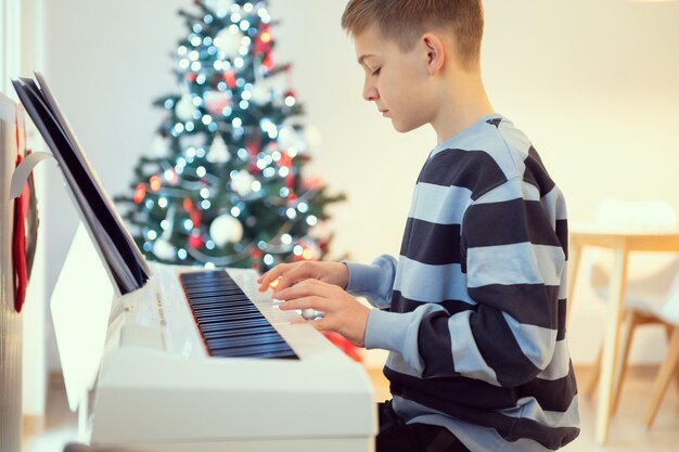 Menino Tennage praticando piano em casa com árvore de ano novo no fundo