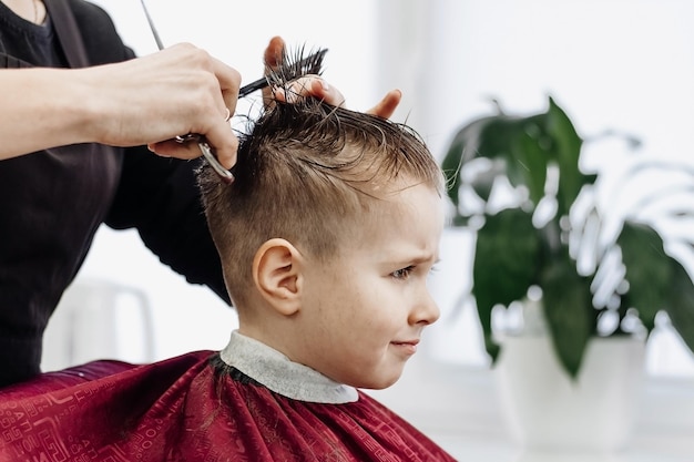 Menino triste corta o cabelo com uma tesoura de barbeiro Closeup de mãos de mulher preparando o cabelo de menino na barbearia Retrato de criança do sexo masculino na barbearia para cortar o cabelo