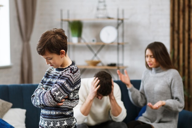 Menino triste fica chateado enquanto seus pais brigam no fundo. Mãe e pai brigam. Conflitos familiares e divórcio levam a problemas psicológicos dos filhos. Transtornos mentais em crianças.