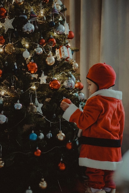 Menino vestido de Papai Noel de pé junto à árvore de Natal