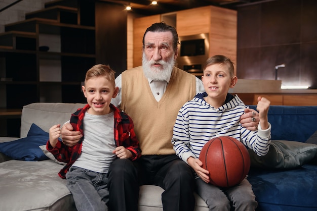 Meninos no sofá com o avô torcendo por um jogo de basquete e segurando uma bola de basquete. Fanáticos esportivos enormes.