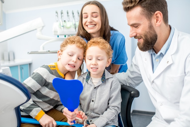 Meninos olhando para o espelho com um sorriso cheio de dentes, sentados com o dentista e a assistente no consultório odontológico