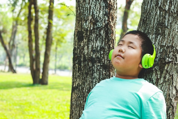 Meninos que usam fones de ouvido verdes da música que estão contra a árvore, olhos fechados, felizes.