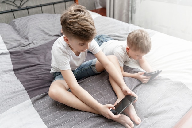 Meninos tristes irmãos estão sentados na cama com telefones nas mãos.