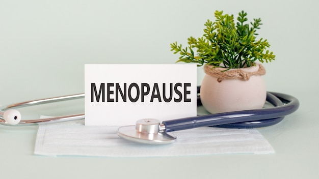 MENOPAUSIA palabras escritas en tarjeta médica blanca, con máscara de medicina, estetoscopio y flor verde en la pared