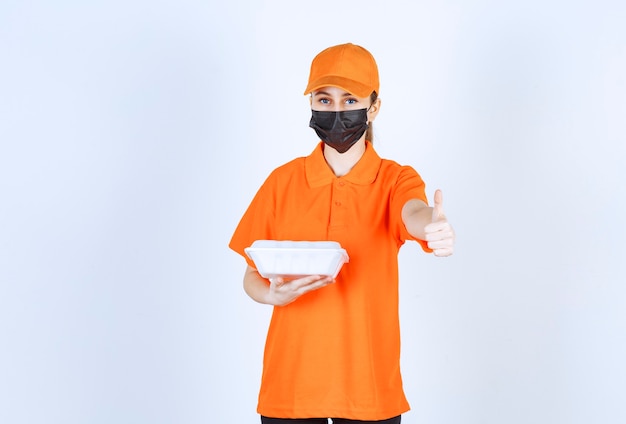 Mensageira de uniforme laranja e máscara preta segurando uma caixa de comida para viagem de plástico e apreciando o sabor