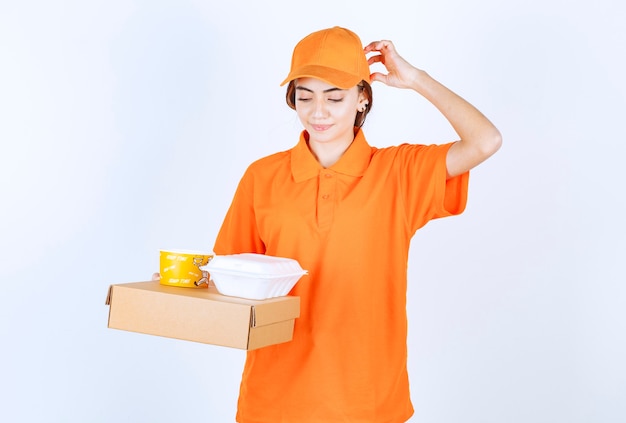 Mensageira feminina com uniforme laranja segurando caixas amarelas e brancas para viagem com um pacote de papelão e parece confusa e pensativa
