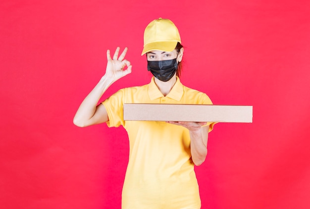 Mensageira feminina em uniforme amarelo e máscara preta segurando a caixa de papelão e mostrando sinal positivo com a mão