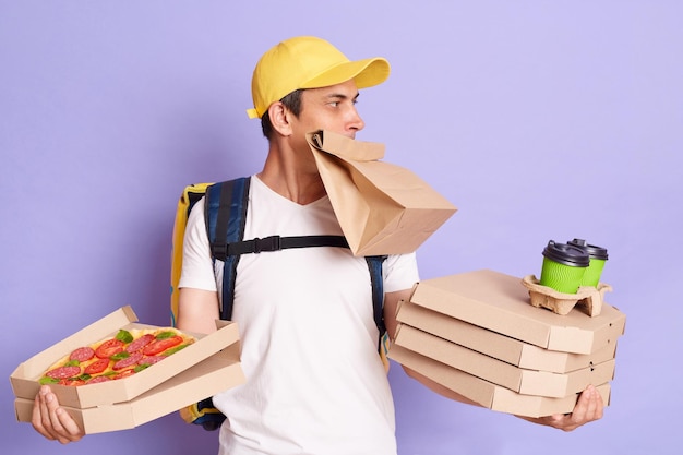 Mensageiro vestindo camiseta e boné segurando caixas de pizza e café para viagem nas mãos segura pacote de papel na boca olhando de lado tem muitos pedidos posando isolados sobre fundo roxo