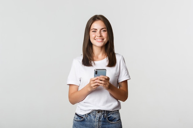 Mensagem de mensagens de texto atraente sorridente menina morena, usando telefone celular e olhando para a câmera.