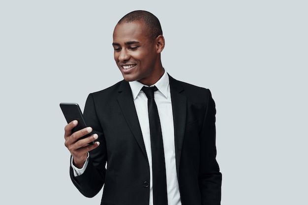 Mensagem de texto. jovem africano charmoso em trajes formais, usando um telefone inteligente e sorrindo em pé contra um fundo cinza