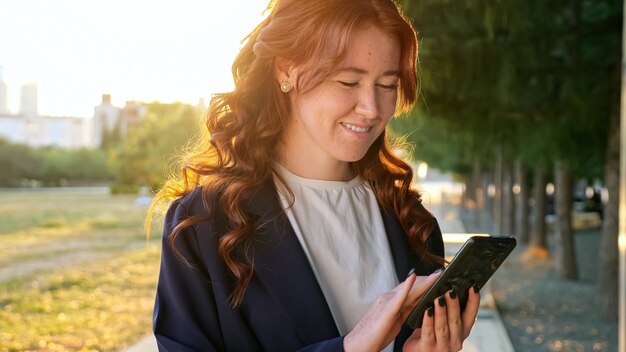 Mensagem de texto sorridente de senhora de cabelo encaracolado de cobre no smartphone moderno em pé na estrada do parque contra uma cidade distante na luz do sol vista de perto