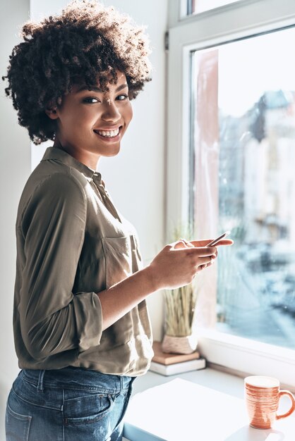 Mensaje de un amigo. Atractiva joven africana usando su teléfono inteligente y sonriendo mientras está de pie cerca de la ventana en el interior